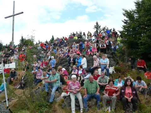 Viele Besucher sitzen auf dem Felsgipfel rund um ein hohes Gipfelkreuz
